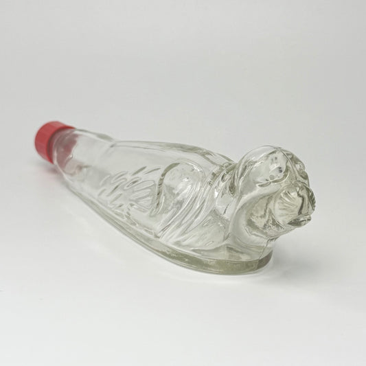 Vintage tysk figurflaske i klart formpresset glas, udformet som en liggende sæl – Seehund.
Flasken har detaljeret relief med sælens hoved, luffer og krop. Hoved og snude er udført i massivt glas.
Denne type figurflaske blev fremstillet i Tyskland som emballage til spiritus. Tilsvarende eksemplarer ses med originale etiketter mærket bl.a. “Seehund” og “Deutsches Erzeugnis”. Producenten af dette eksemplar er ikke identificeret med sikkerhed.
Med original/lignende ældre rød skrueprop i plast med kork på ind